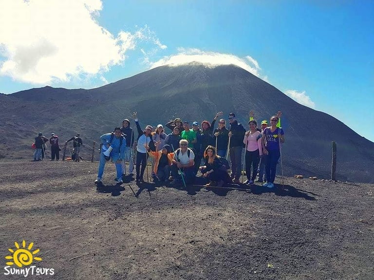 Group of tourists posing together on dark volcanic terrain with a large snow-capped volcano in the background under blue sky