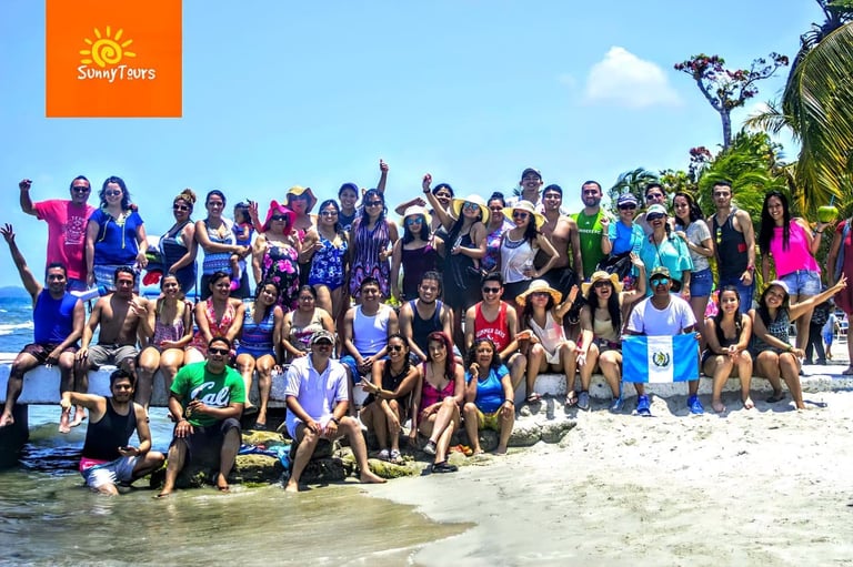 Large group of diverse people posing together on a sandy beach in shallow water, wearing colorful clothes and wetsuits for water activities