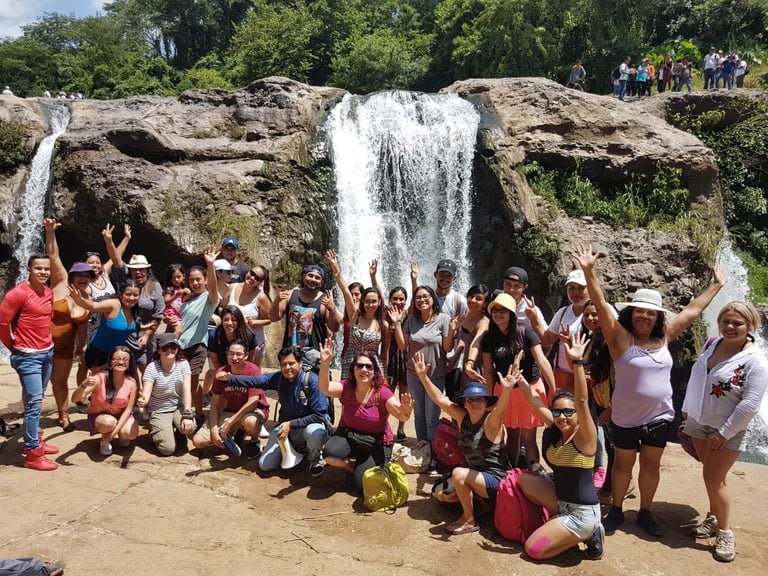 Large group of people posing together in front of a waterfall with their arms raised