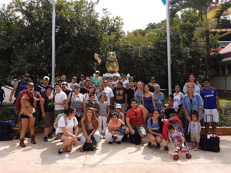 Large group of diverse people posing together outdoors in front of a yellow statue, surrounded by trees and palm trees