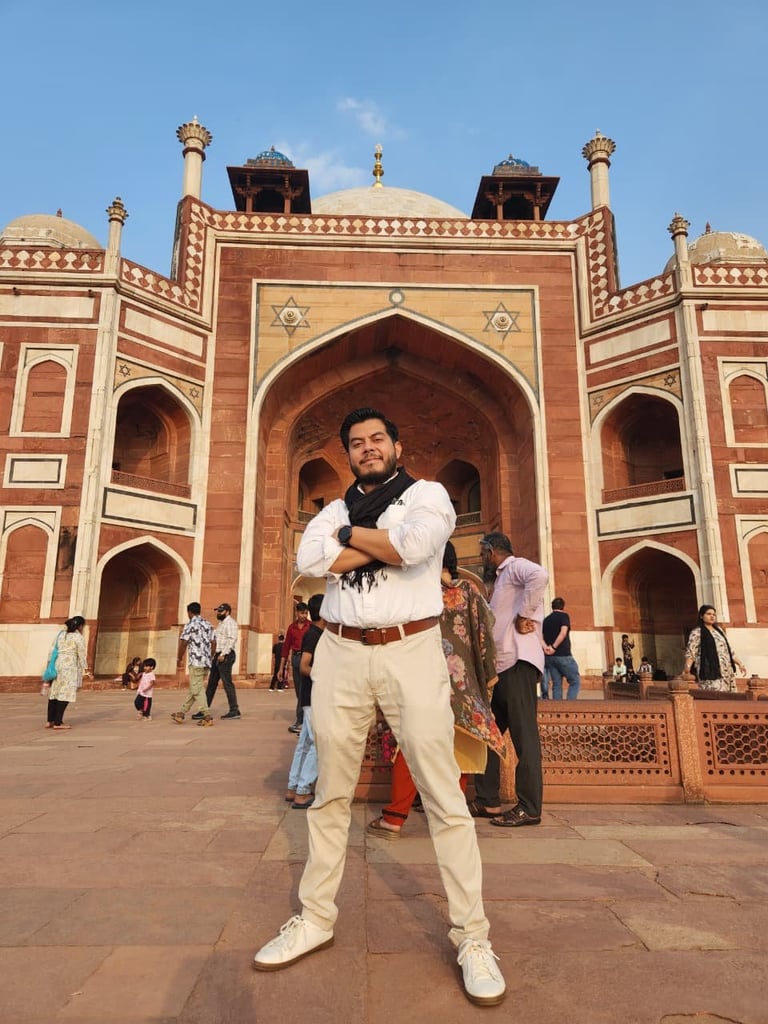 Man posing in front of red brick Mughal mosque with white shirts and cream pants, tourists visible in background courtyard